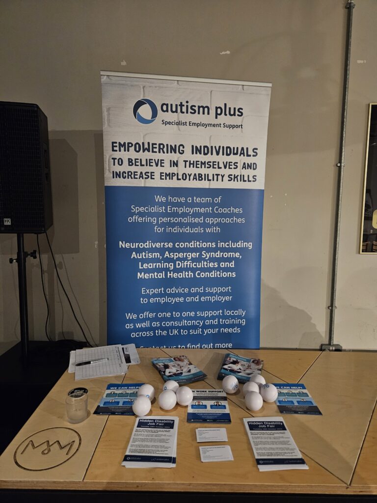 A picture of a marketing banner for Autism Plus Employment services behind a table that has leaflets on and some stress ball marketing items